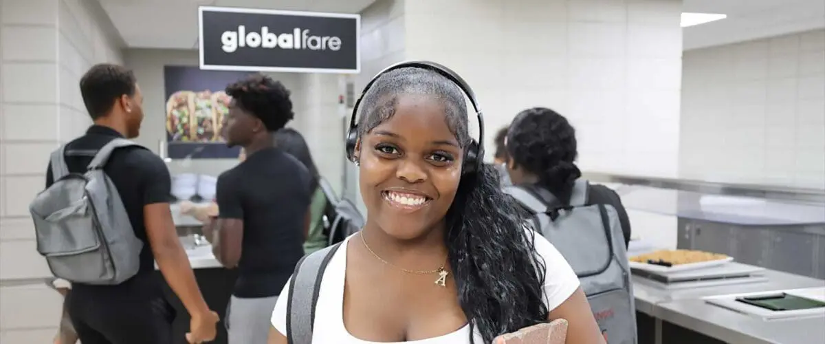 Student smiling while holding books and plate of food in the serving line of cafeteria