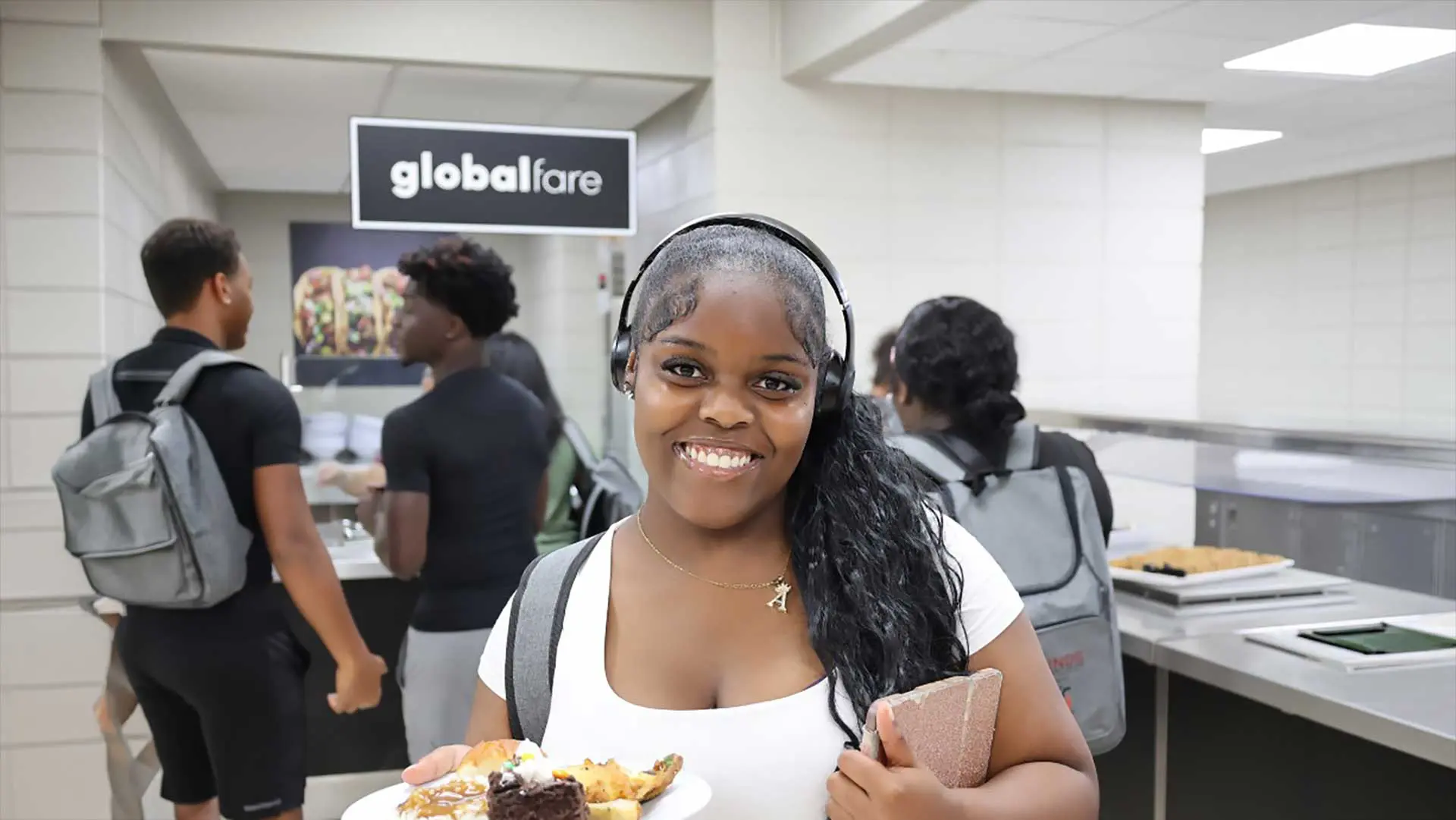 Student smiling while holding books and plate of food in the serving line of cafeteria